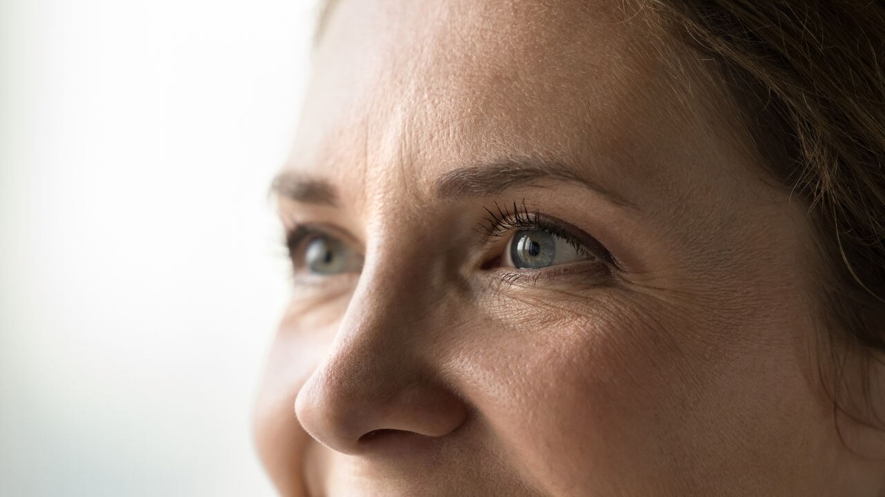 Blue eyes of young middle aged woman with black mascara on eyelashes looking away, smiling, laughing. Cropped shot banner. Female model upper face with natural makeup, dry facial skin with wrinkles