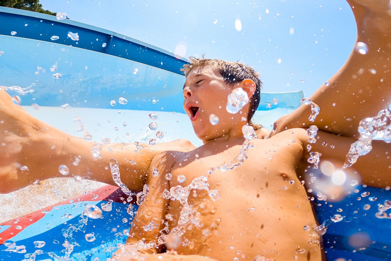 Child with eyes closed splashed with drops of water while is sliding fast through a water slide at a Aqua park during a hot day of summer vacation. Happiness in holidays.