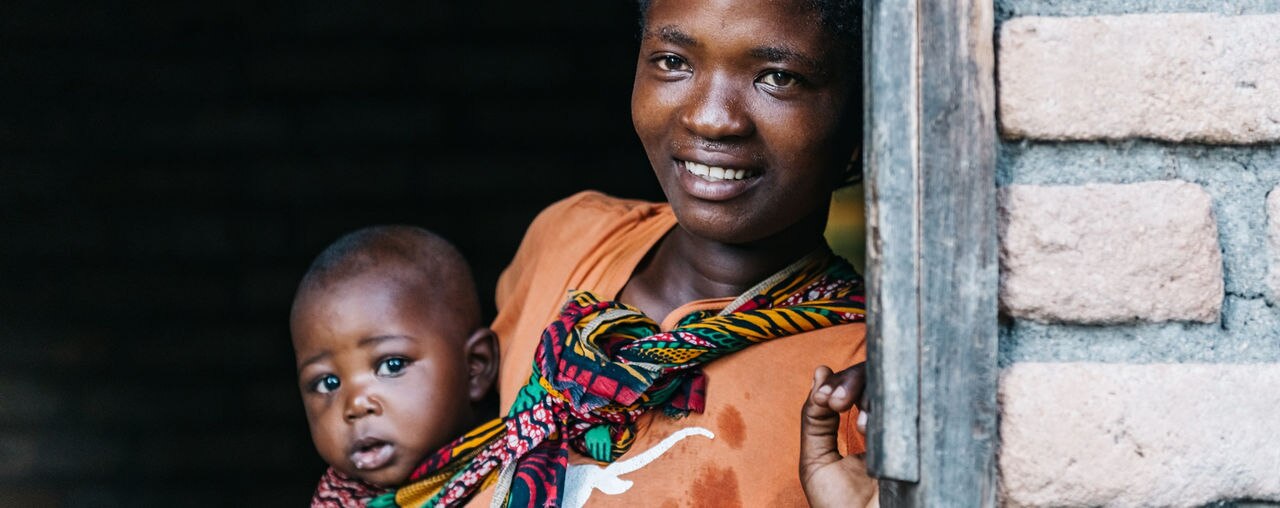 young african mother with baby looking out of the door of typical brick house