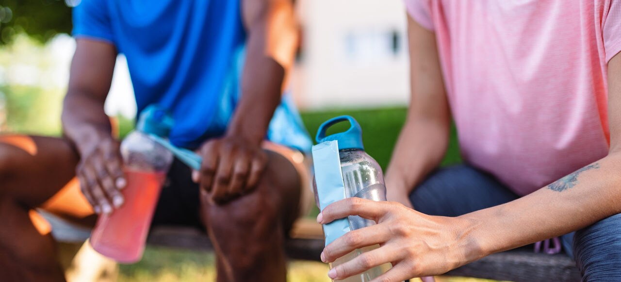 Two people sit outdoors on a bench, hydrating with water bottles after exercising. The image showcases fitness, relaxation, and a healthy, active lifestyle.