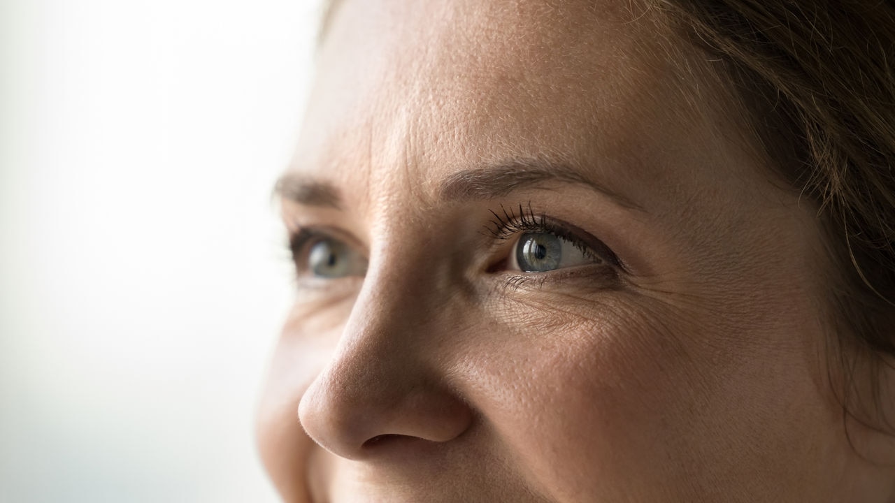 Blue eyes of young middle aged woman with black mascara on eyelashes looking away, smiling, laughing. Cropped shot banner. Female model upper face with natural makeup, dry facial skin with wrinkles