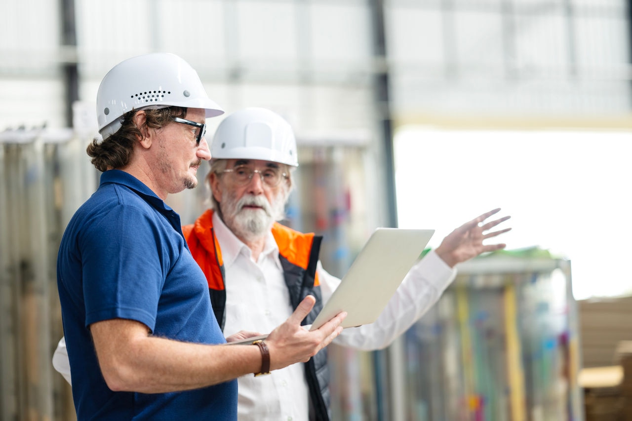 Running Your Manufacturing Operation Smoothy with the Manufacturing Operations Management System Optimizer. Business Partner having a discussion over a laptop computer on the production line factory floor to increase production efficiency and reduce time.