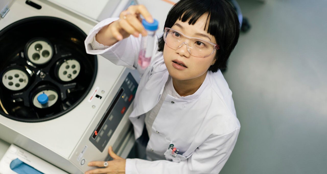 A asian female scientist is working inside a laboratory. She's taking a close look and examining a flask that cotains a pinkish liquid.