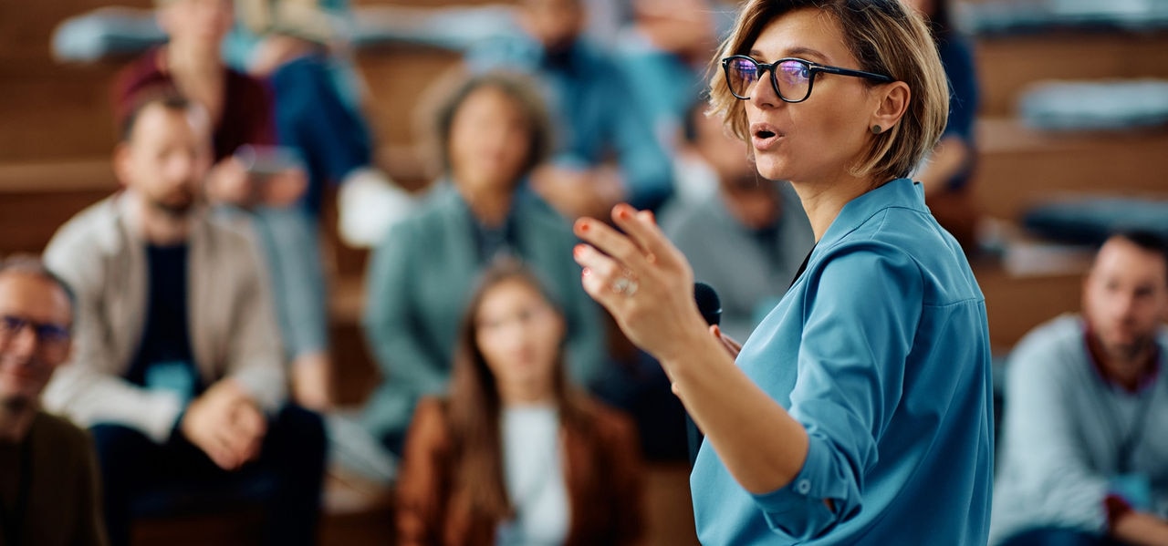 Mid adult businesswoman talking to group of seminar attendees in conference hall.