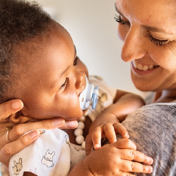 Smiling mom holding her adorable little adopted baby girl with a pacifier in her mouth in her arms at home