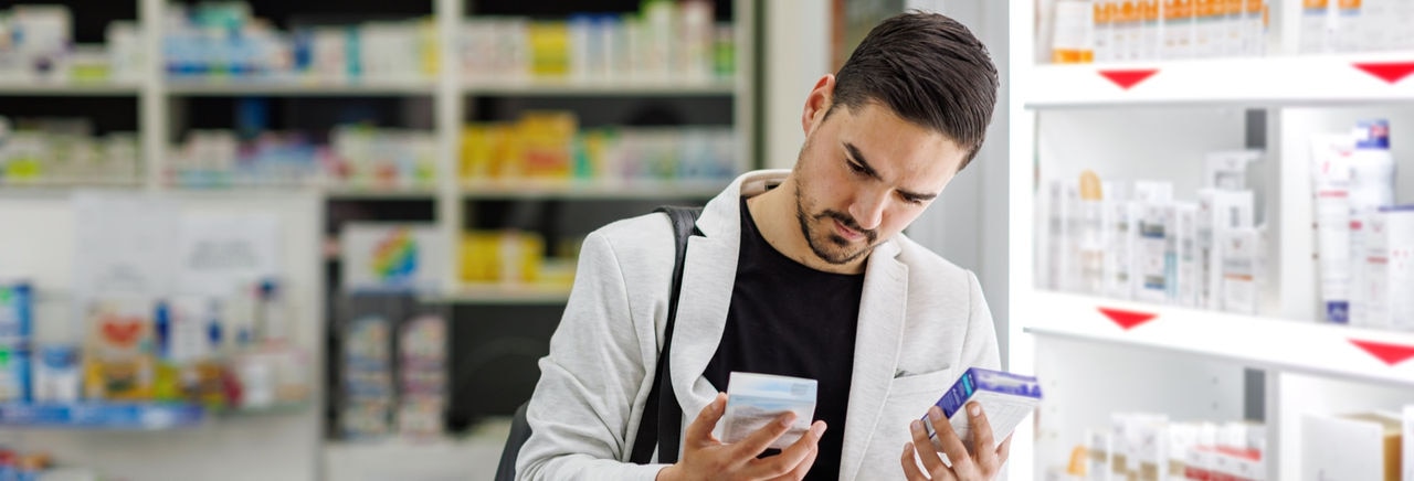 A young Caucasian businessman is standing in a pharmacy and choosing betweeen two boxes of pills.