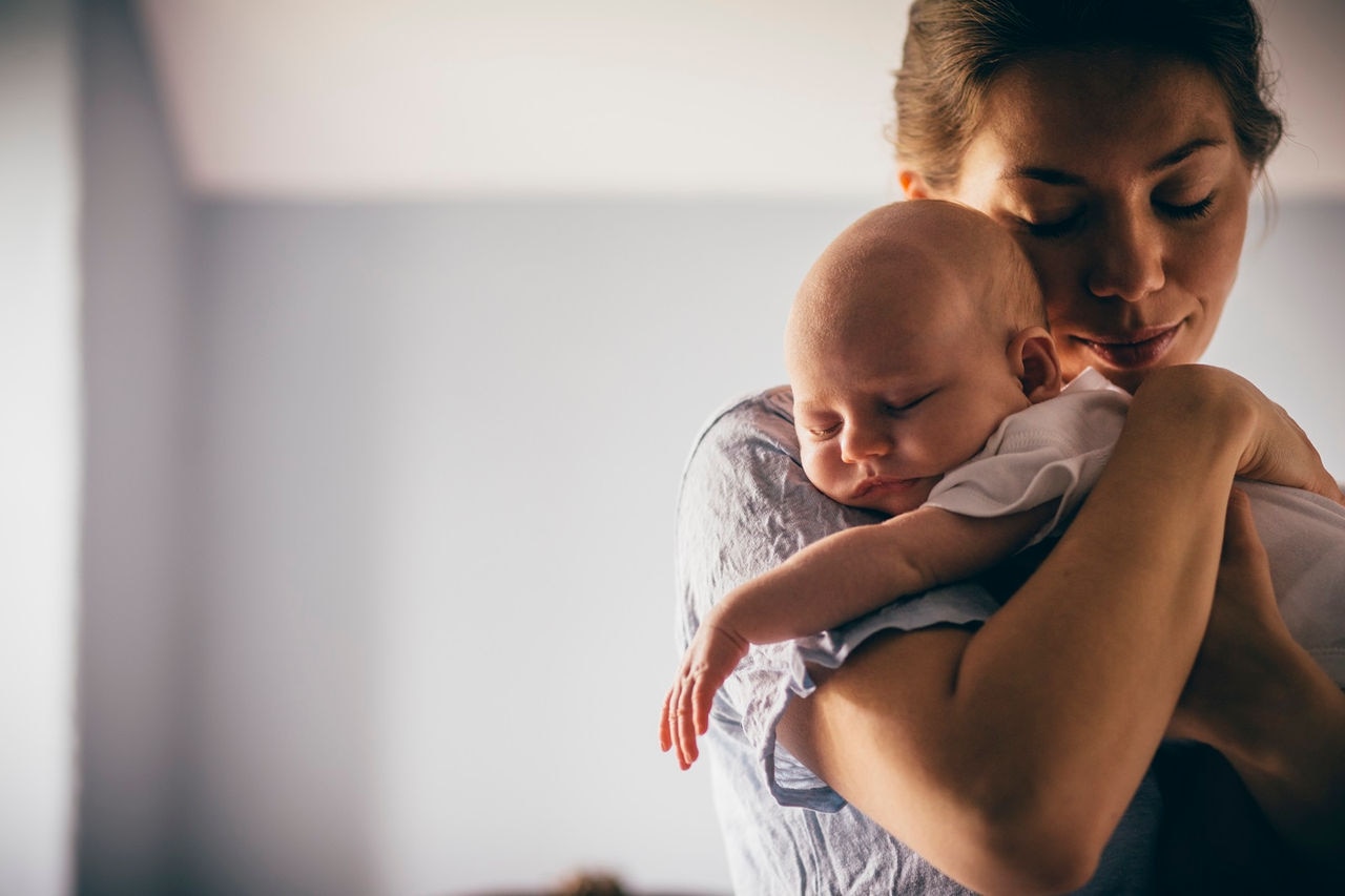 A happy new mother smiles as she craddles her sleeping baby in her arms. She has her eyes closed as she holds her close.