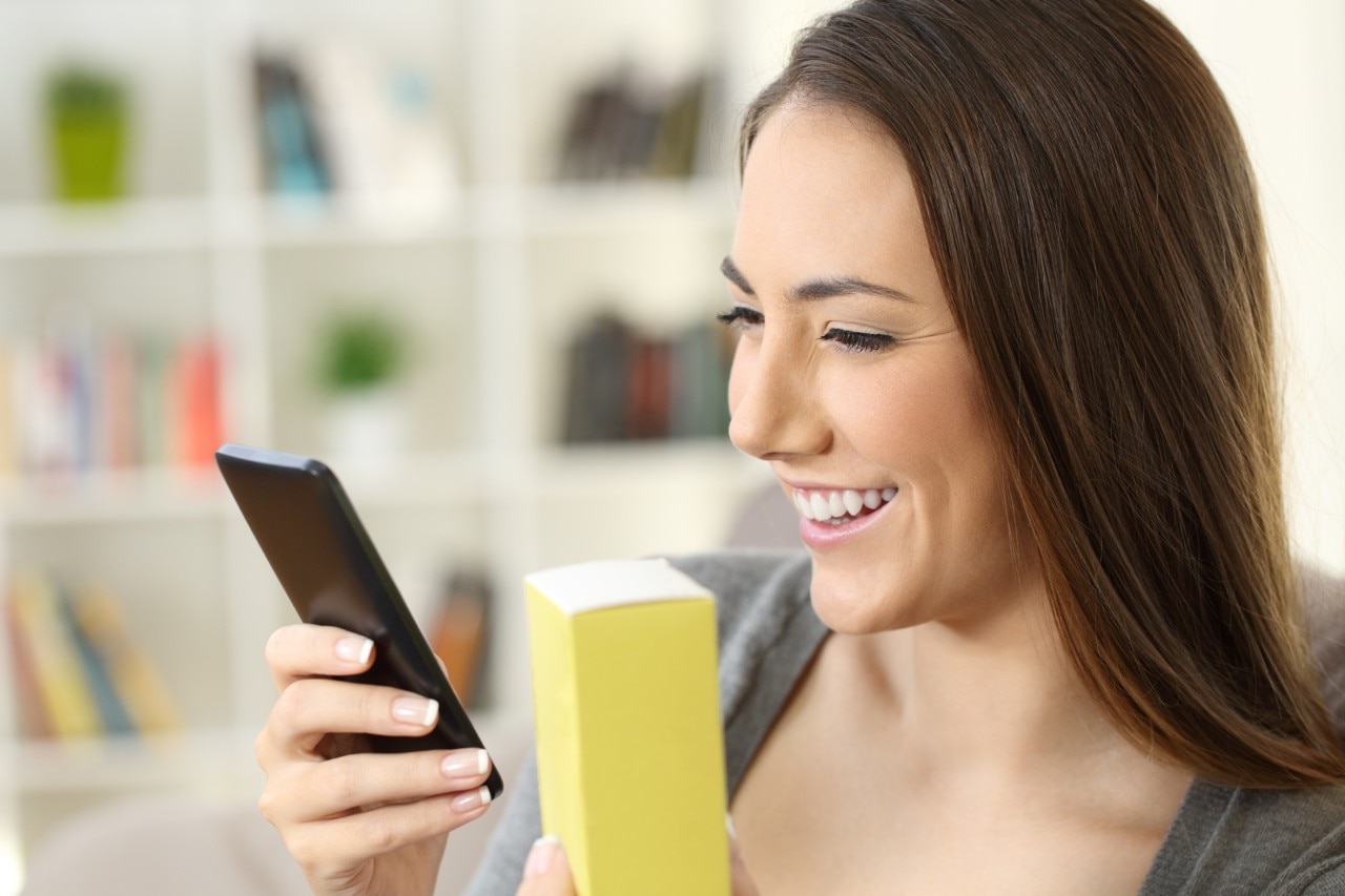 Happy girl reading on line information on a smart phone about a medicament box sitting on a couch in a house interior