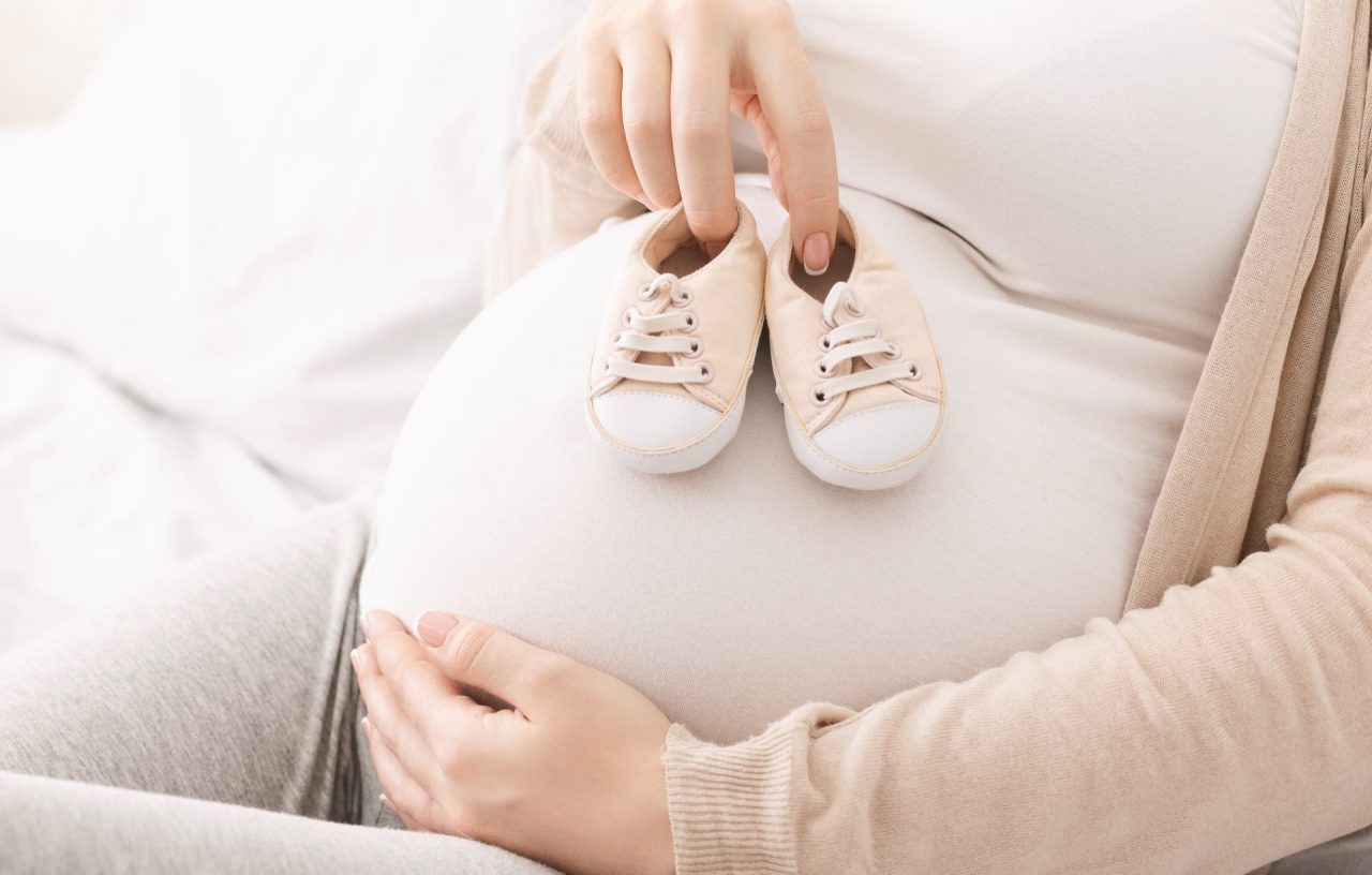 Millennial woman playing with her belly, trying tiny baby shoes on it, close up, panorama with free space