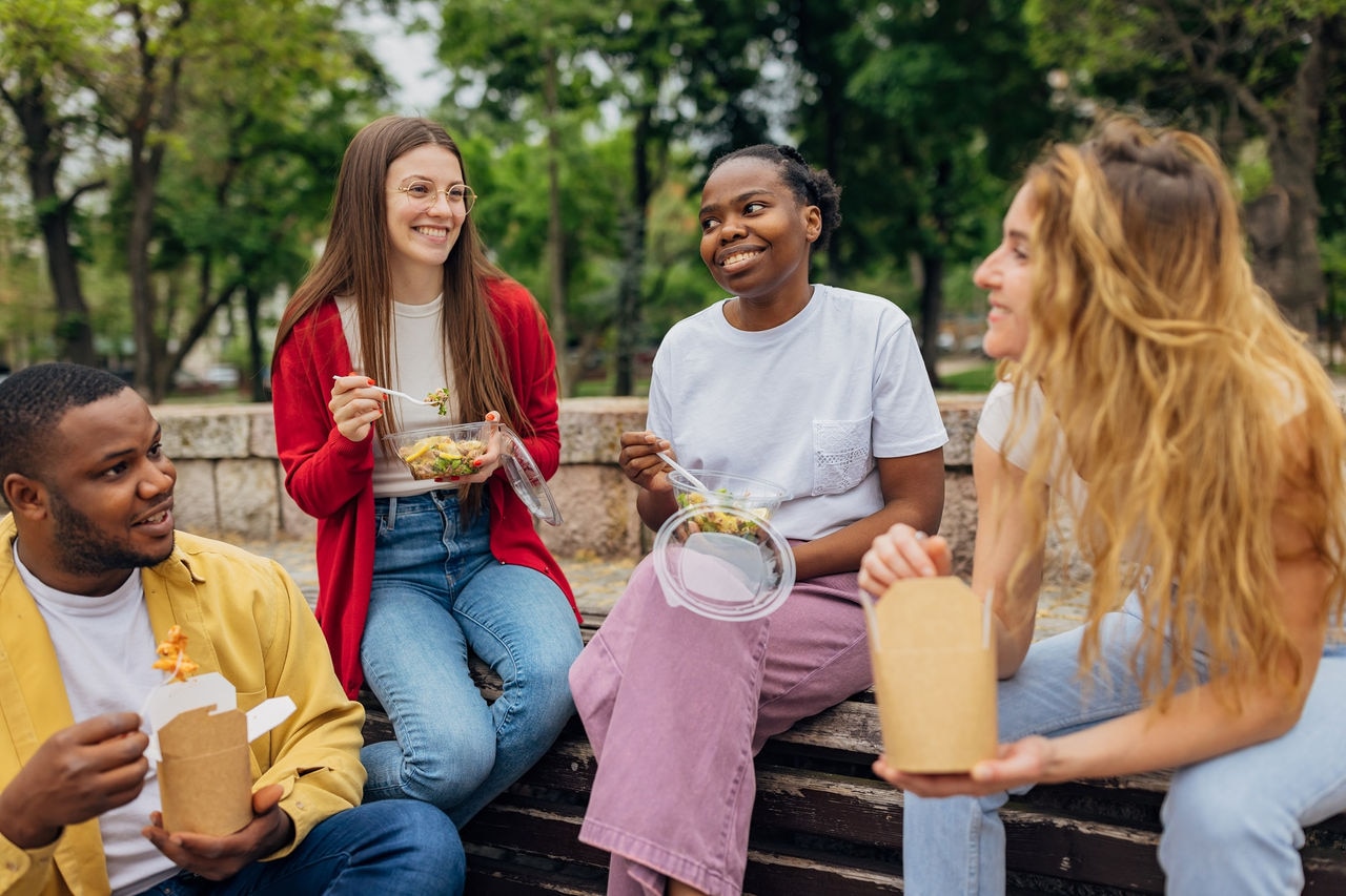 Small group of diverse friends having fun eating healthy take out food in the park