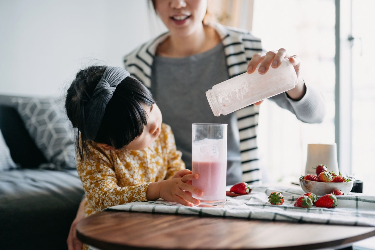 Young Asian mother making healthy strawberry smoothie with fresh fruit with lovely little daughter at home. Little daughter is excited about the drink while mother is pouring to glass. Healthy eating and healthy lifestyle