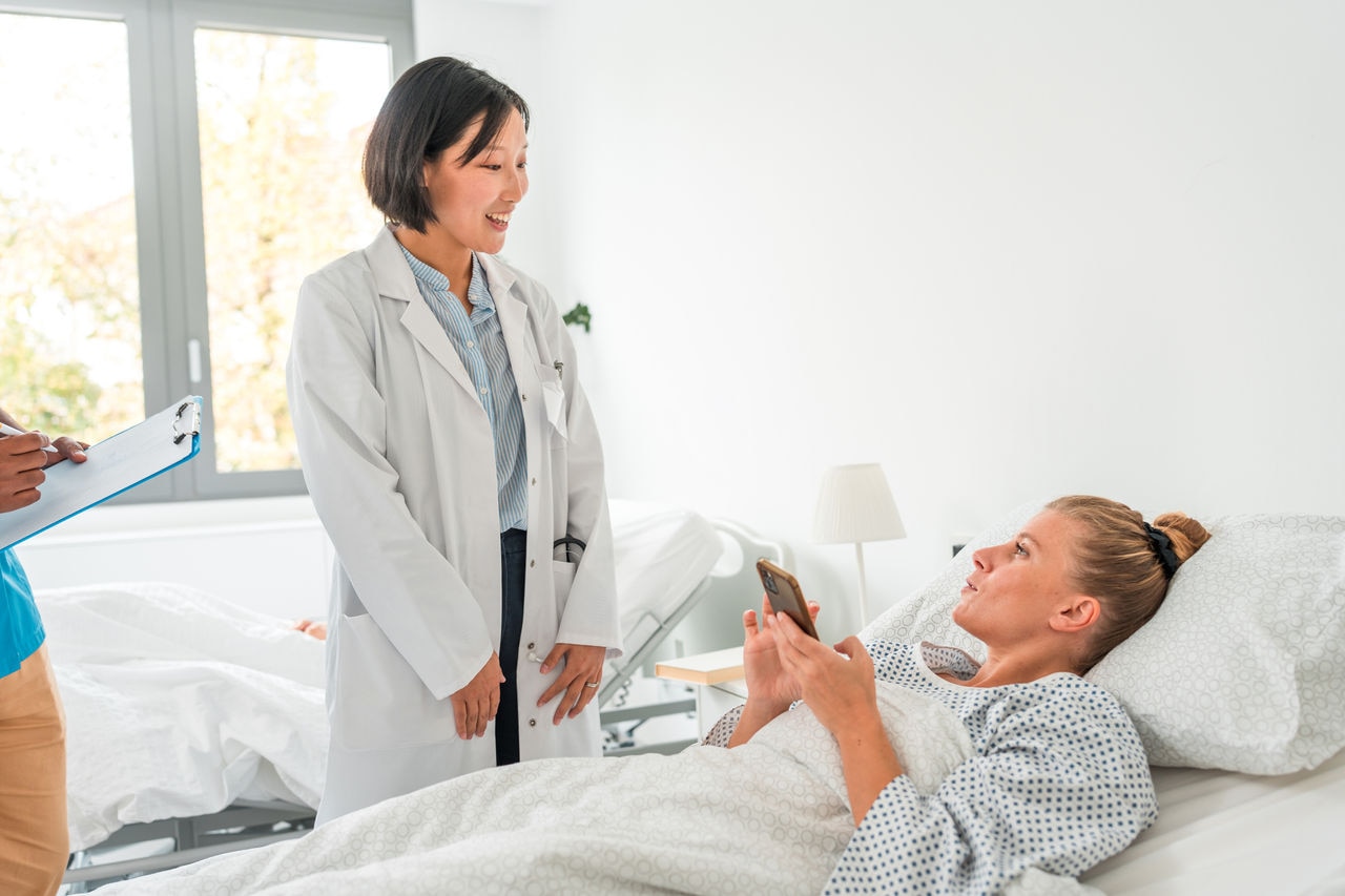 Mid-adult Asian female doctor performs a routine check on a female Caucasian patient lying in a hospital bed during recovery.