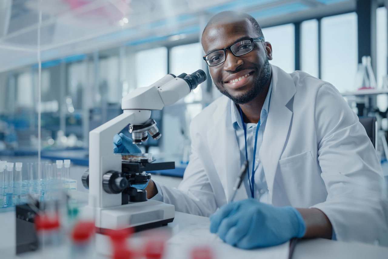 Modern Medical Research Laboratory: Portrait of Male Scientist Using Microscope, Charmingly Smiling on Camera. Advanced Scientific Lab for Medicine, Biotechnology, Microbiology Development