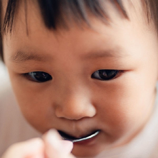 Close-up, over the shoulder view of Asian little girl eating food, fed by her mum with a spoon. Caring and togetherness concept.