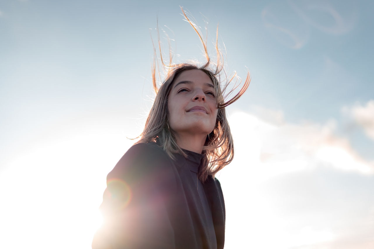 A young woman is captured from the waist up, smiling and enjoying the breeze and sunset from a mountain viewpoint. Her radiant smile reflects the joy she feels as the wind gently tousles her hair, and the sun casts a warm, golden glow over the majestic mountains in the background.