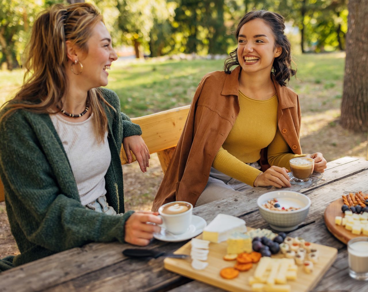 Two smiling female friends sit on the bench at a wooden table in nature and enjoy chatting and snacking on crackers and different types of cheese