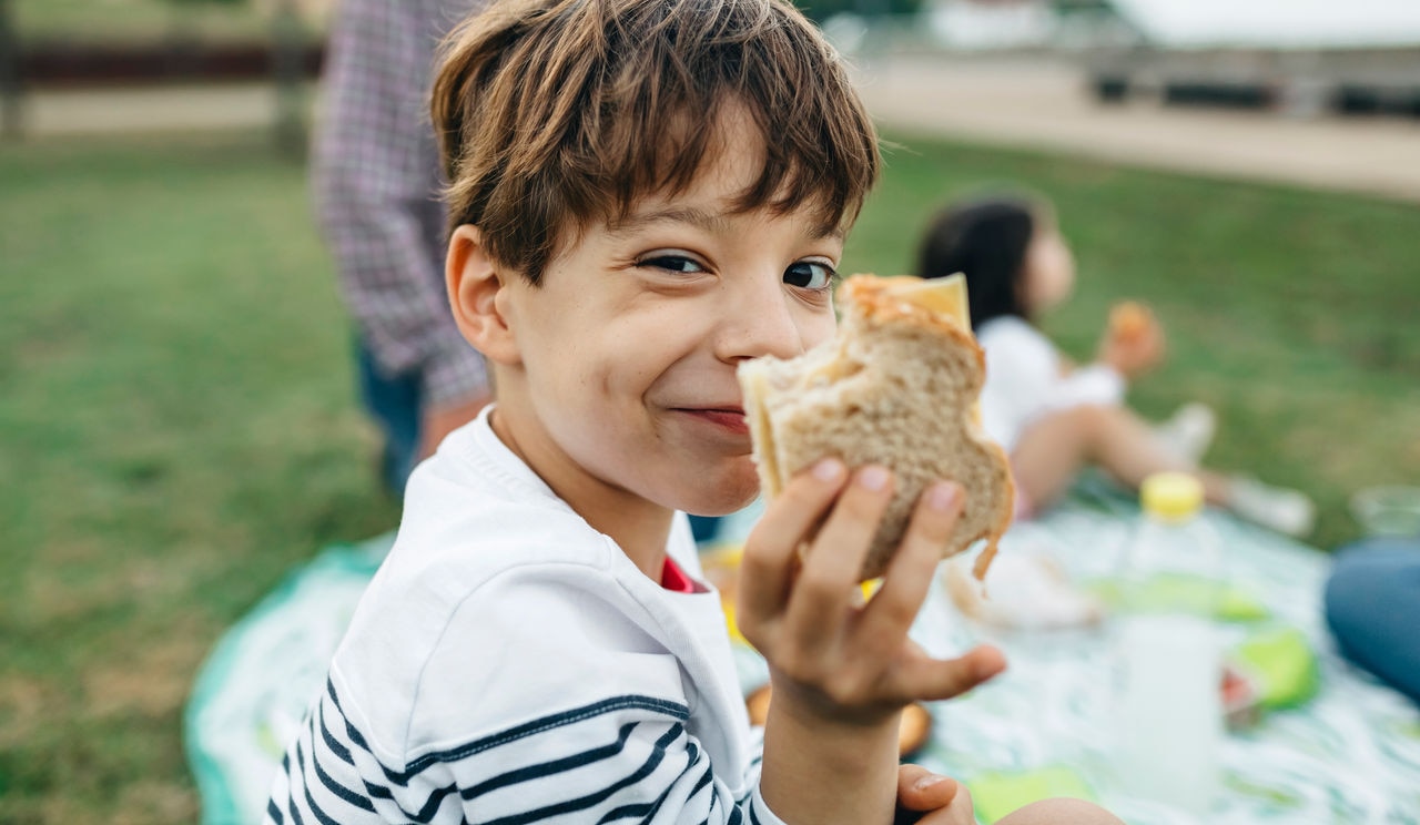 Spain, Asturias, Close up of happy kid looking at camera while showing a homemade sandwich with his family eating over the grass on background