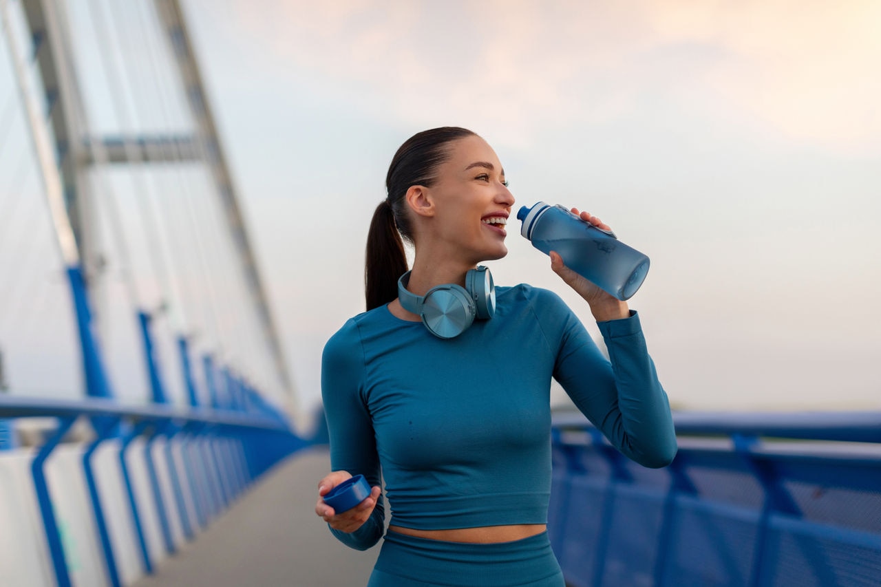Excited young woman taking break from jogging, having break and drinking water, holding bottle to refresh herself during outdoor workout in the evening