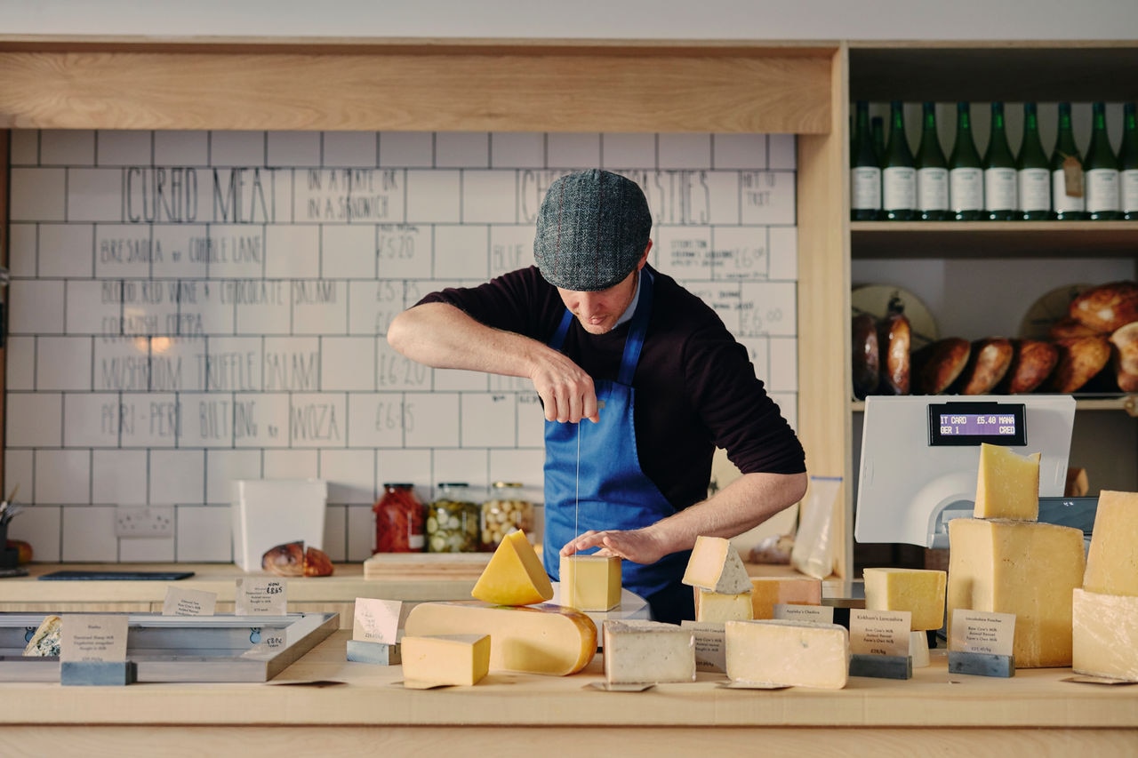 Cheesemonger cutting cheese with cheese wire surrounded by a variety of cheese on counter top