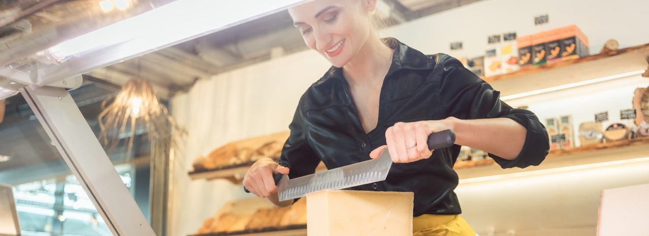 Young shop clerk in deli cutting cheese at the counter with a knife