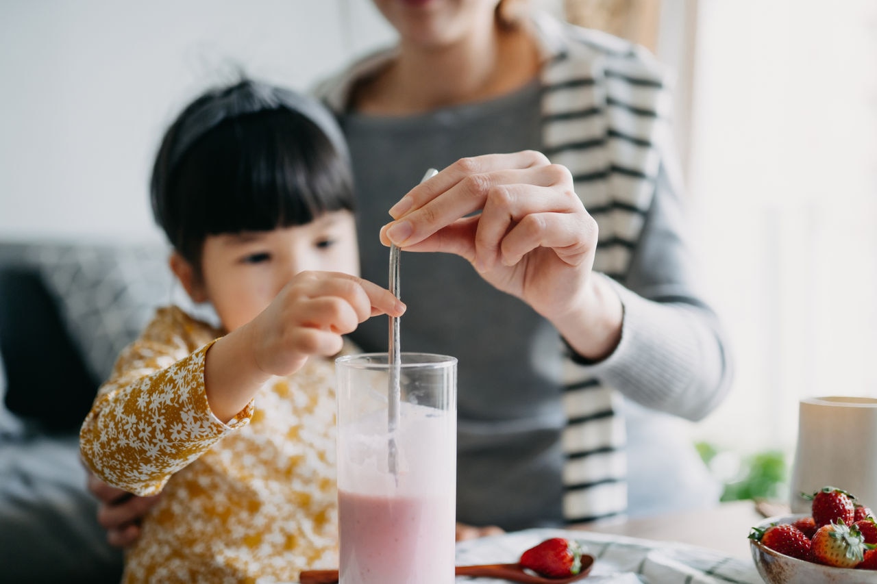 Young Asian mother making healthy strawberry smoothie with fresh fruit with lovely little daughter at home. Mother and daughter drinking the juice with a reusable metal straw. Sustainable lifestyle, healthy eating and healthy lifestyle