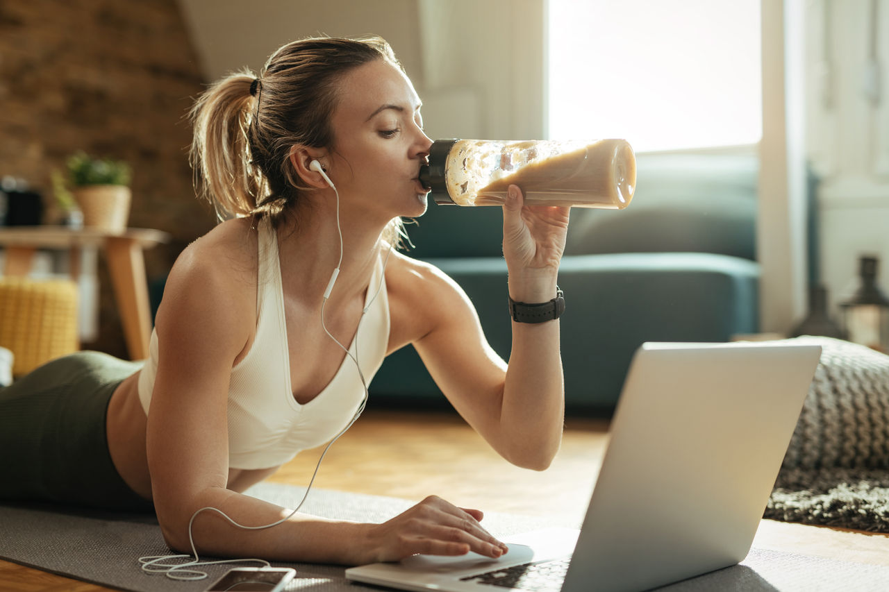 Young sportswoman relaxing on the floor and drinking smoothie while using laptop at home. 