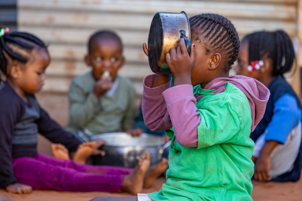 Poverty in Africa. Young starving Black African girl eating maize from a pot, three children in the background