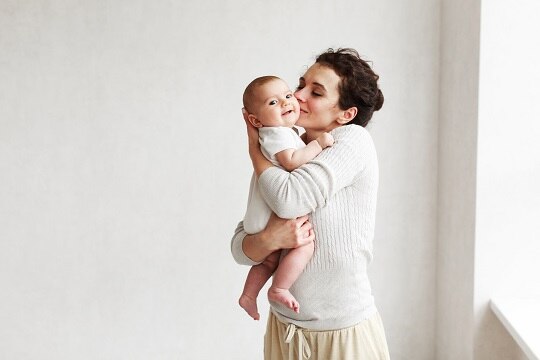 Beautiful caucasian middleaged woman with dark hair in home cozy clothes at home by the window holding a three-month-old baby on a white wall background