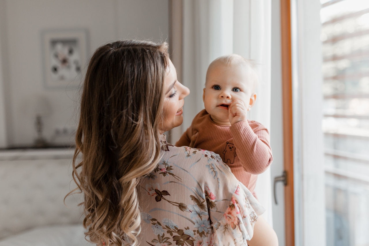 Young woman mom with baby girl on hands near window at home