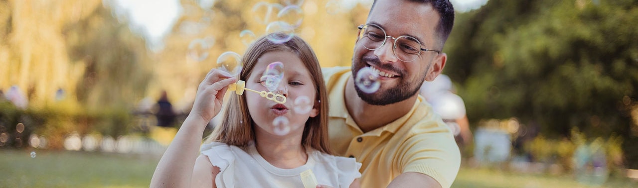 Outdoor Portrait of Little Daughter With Her Father Making Bubbles in the Public Garden at Vacation Day or Holiday. Family Concept.