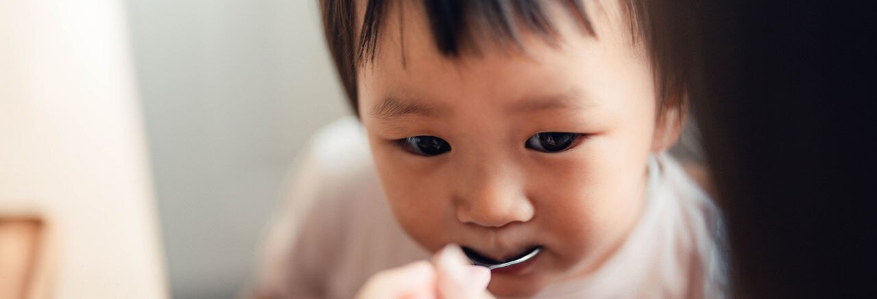 Close-up, over the shoulder view of Asian little girl eating food, fed by her mum with a spoon. Caring and togetherness concept.