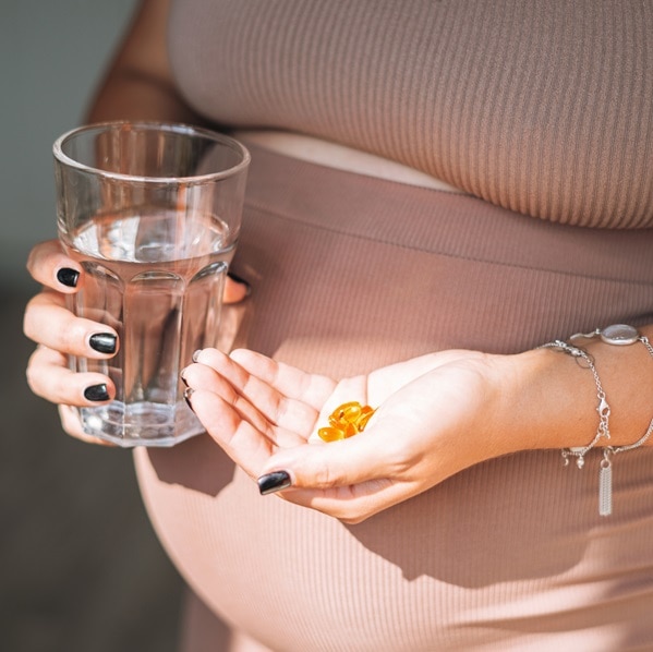 Crop photo of young pregnant woman with glass of water and omega 3 dietary supplements in her hands in kitchen at home, pregnancy health and wellness