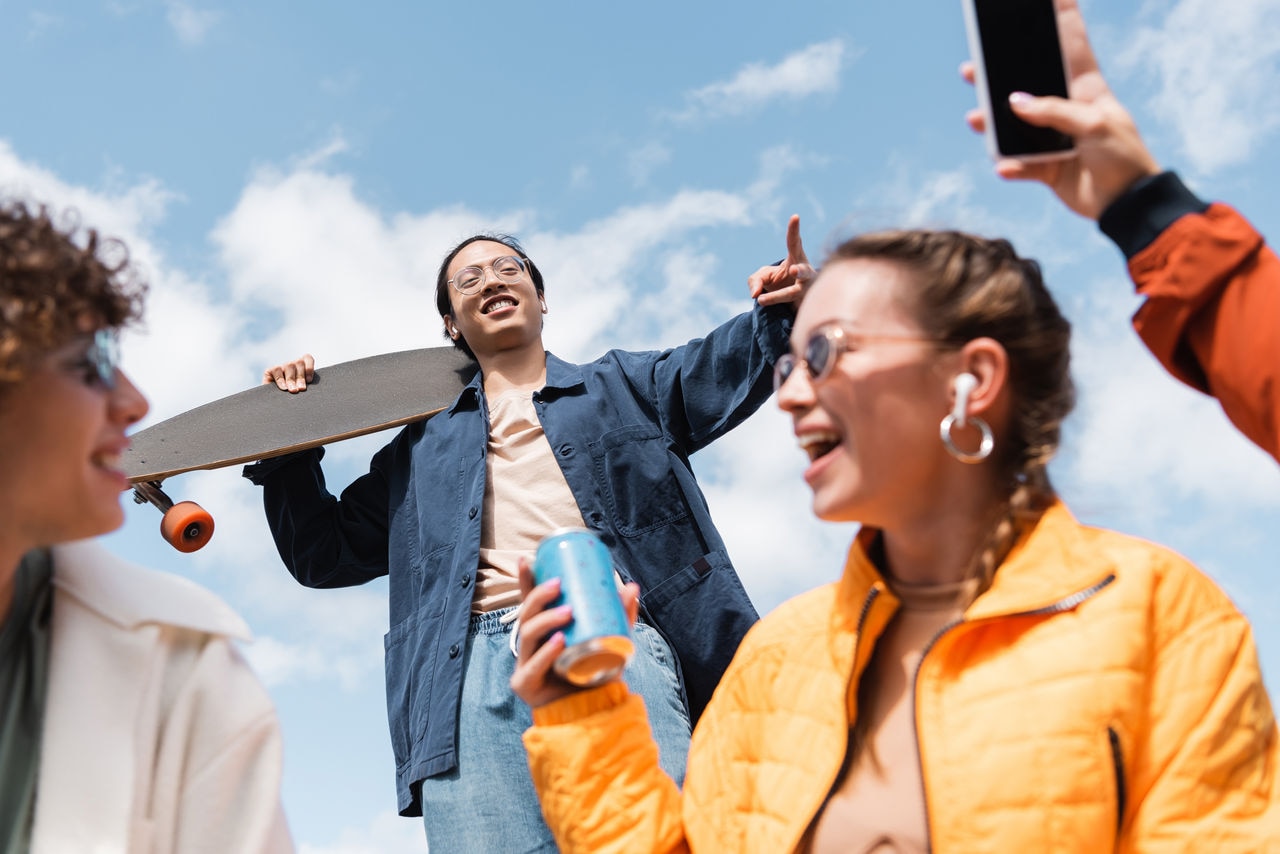 low angle view of asian man with longboard showing victory sign near happy friends