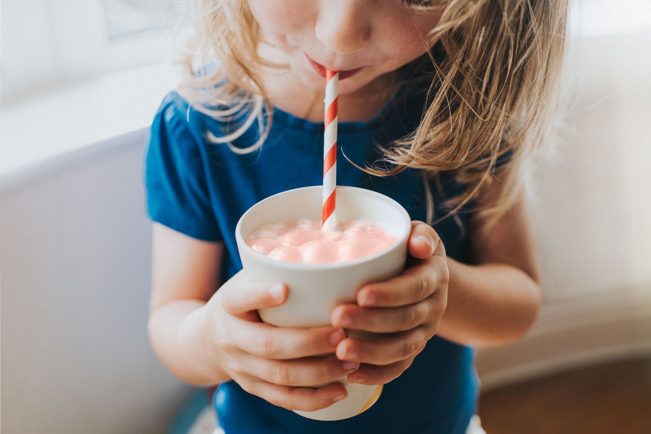 Cute little pre-school age girl gripping a cup of milk and sucking from a red striped straw. Space for copy.