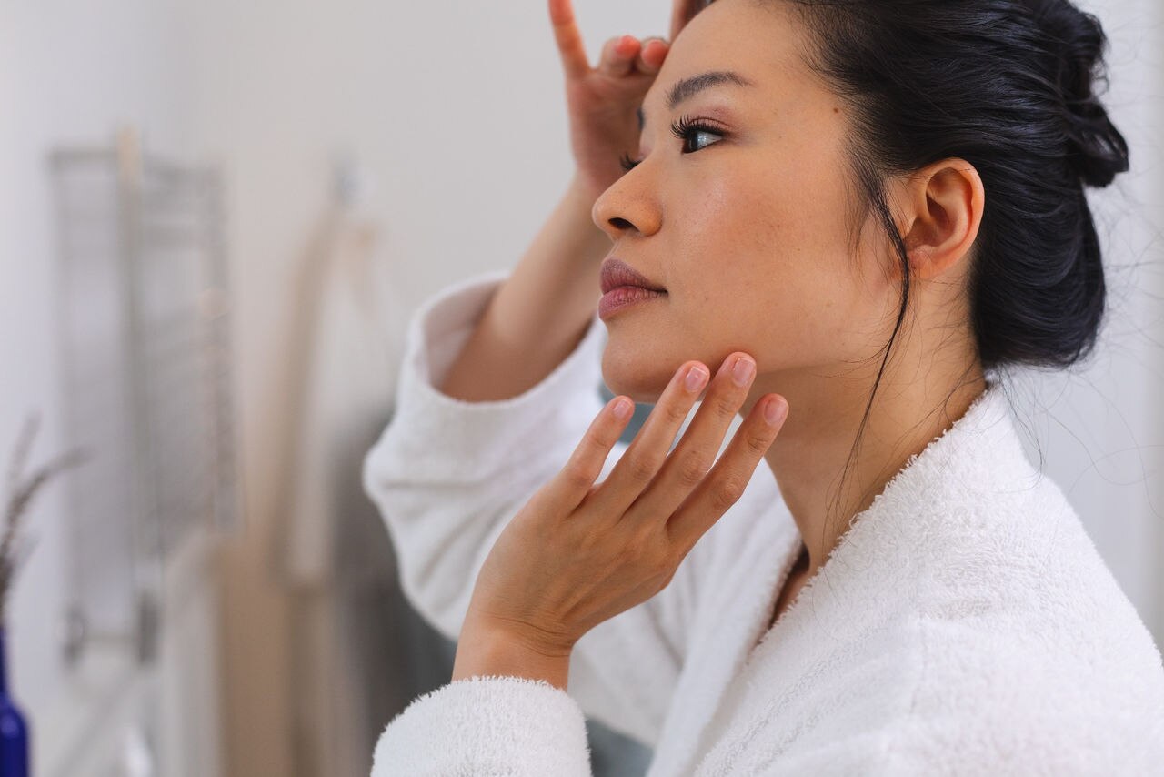 Happy asian woman wearing bathrobe standing in front of mirror applying face cream in bathroom. lifestyle, self care, health and beauty.; Shutterstock ID 2285537035; purchase_order: P&B Loop 1&2(26)-1*.16.01.2024; job: ; client: ; other: 
