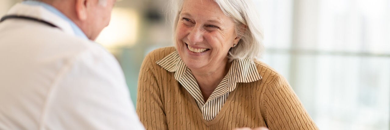 A senior female patient is meeting with her doctor. The two are seated at a table together in a medical clinic. The patient is smiling warmly at her care provider. The male doctor is talking.