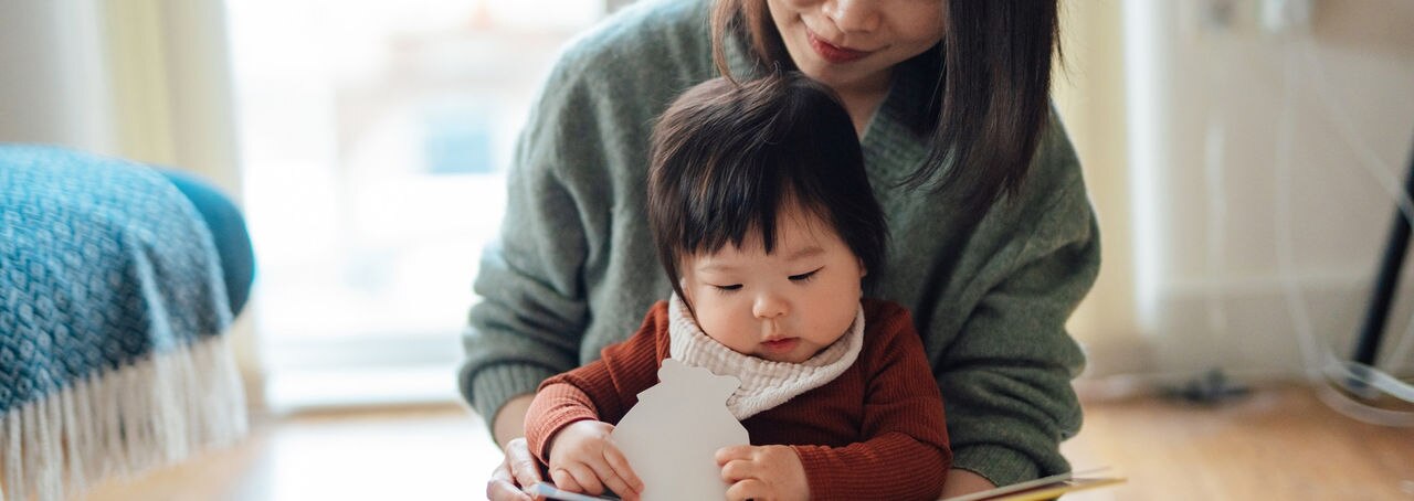 Cute Asian baby girl sitting on her mother’s lap, reading story book in the living room. Children education and learning. Mother bonding with baby.