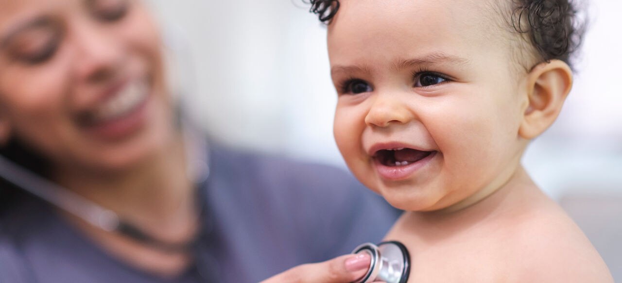 A female doctor of Hispanic descent is doing a routine check-up on a baby. The medical professional is using a stethoscope to check the child’s heart rate. The baby is seated on a medical exam table and is smiling.