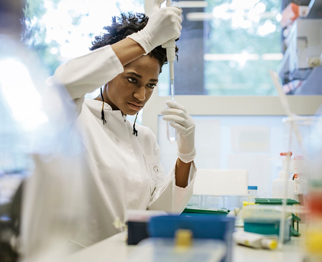 A black female scientist is pipetting in a laboratory. She's holding a pipette in one hand and a small flask in the other one, while wearing white gloves.