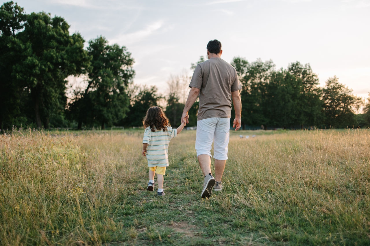 A young dad and his son are walking in the grass field while holding hands.