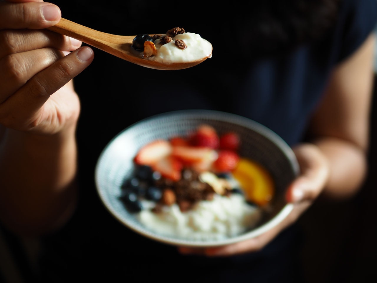 Probiotic and fermented food theme greek yogurt topped with granola and fresh blueberry in wooden spoon with hand holding on blurred background of yogurt bowl with peach, strawberries and blueberries.; Shutterstock ID 1331043386; purchase_order: -; job: -; client: -; other: -