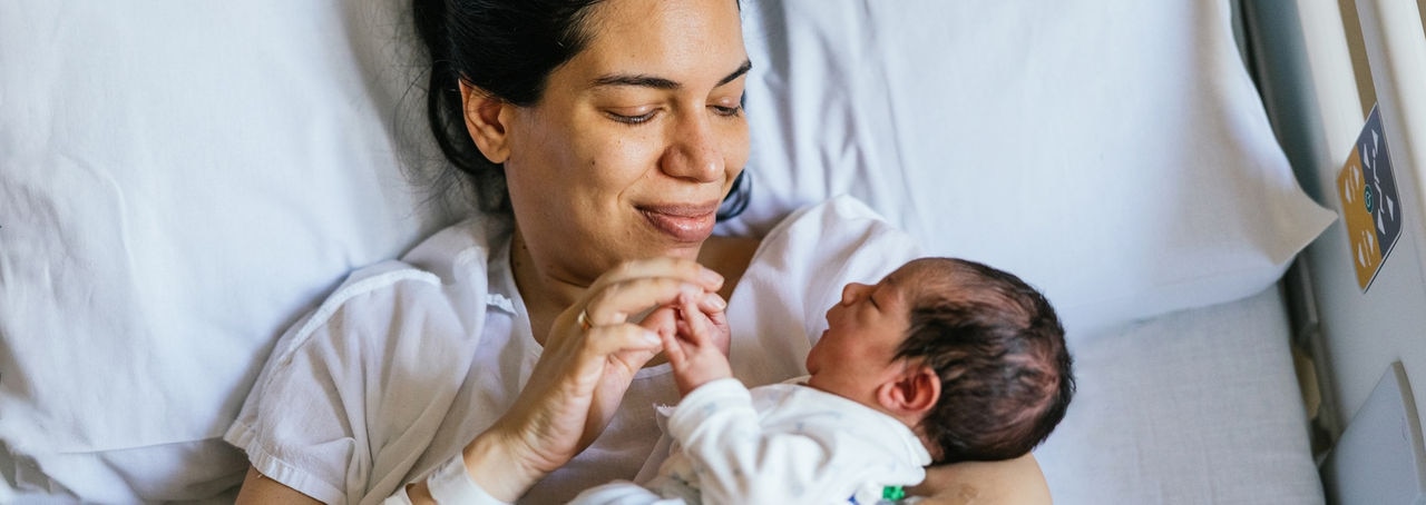 Woman holds the hands of her newborn baby as she rests in a hospital bed. She gazes lovingly at her tiny baby.