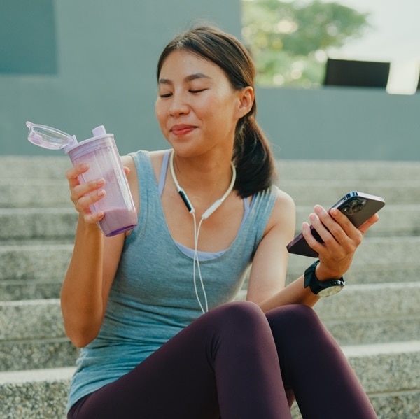 Young Asian female athlete runner drinking protein shake and using smartphone for fitness motivation after exercise workout in urban park. Diet and healthy food workout exercise concept.