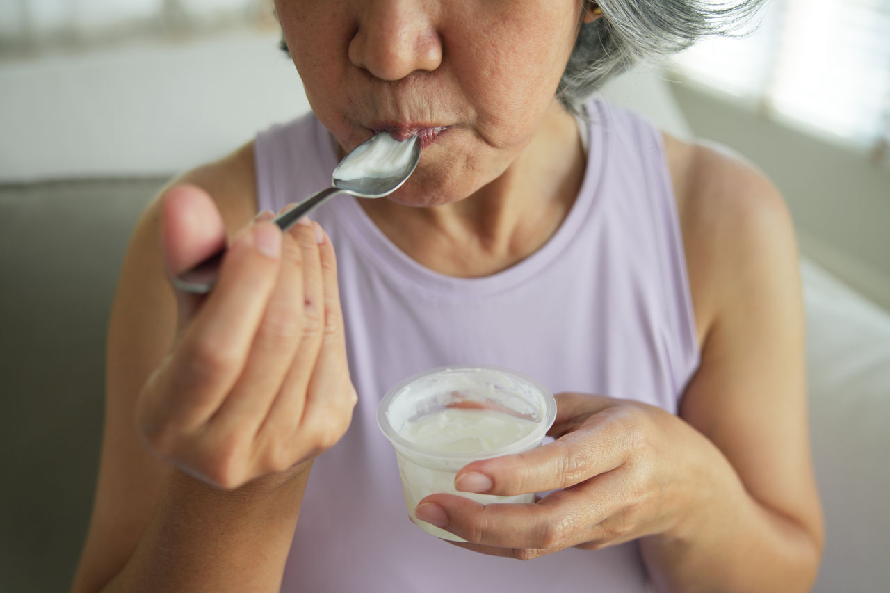 Asia adult people middle age happy old woman eat greek yogurt cup weight loss protein gut health care diet detox sit at home sofa elder aging asian aged female. Fresh medical food relief meal enjoy.