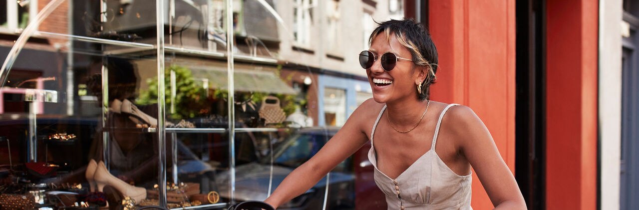 Happy young woman with sunglasses riding bicycle by exclusive 2nd hand vintage store window