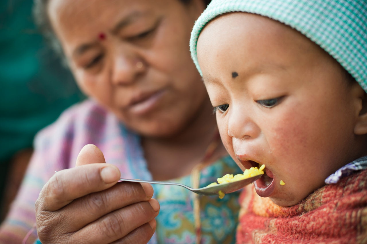 Outdoor image at day time of Asian woman feeding food to her grandson. The little child is opening his mouth wide to eat his lunch fed by his grandmother. Two people, Head and shoulders, horizontal composition and selective focus.