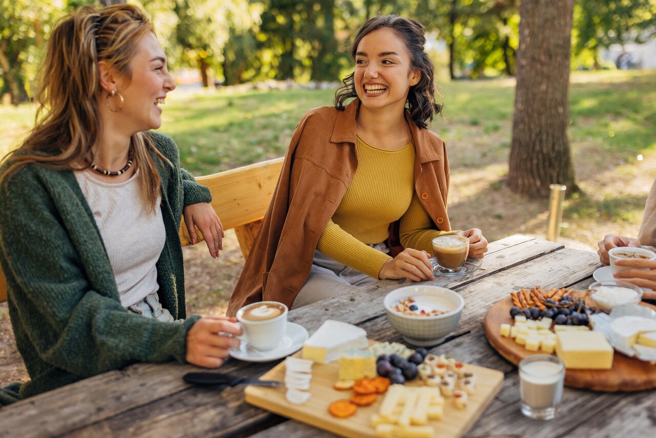 Two smiling female friends sit on the bench at a wooden table in nature and enjoy chatting and snacking on crackers and different types of cheese