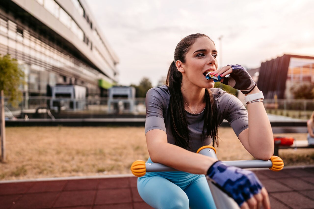 Young beautiful athlete woman eating energy bar after riding outdoor bike in park.