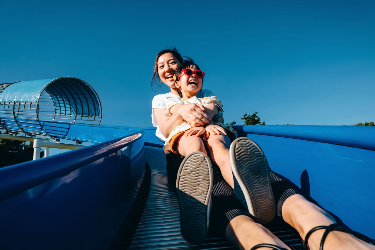Happy young Asian mother with her little daughter having fun playing on the tube slide together in an outdoor playground on a lovely sunny day. Outdoor fun. Happy childhood and motherhood. Mother and daughter bonding moment. Happy family life