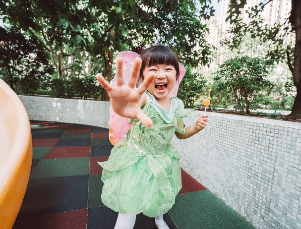 Lovely little girl in a fairy costume reaching out her hand to the camera and playing joyfully while holding a lollipop in her another hand in the playground